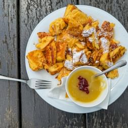 Kaiserschmarrn auf einem weißen Teller mit Puderzucker bestäubt, serviert mit einer kleinen Schale Fruchtsauce und Marmelade auf einem Holztisch.