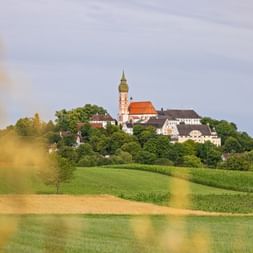 Andechs Monastery with its distinctive church tower sits atop a green hill surrounded by fields and trees under a cloudy sky in Bavaria.