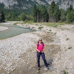 Wanderin in pinkem Shirt mit Rucksack und Stöcken auf steinigem Flussbett der Leutascher Ache, mit Bergen und Wald im Hintergrund.