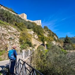 Wanderer mit blauem Rucksack auf Weg zur Festung Rocca Albornoziana in Spoleto, Umbrien. Steinmauern und grüner Hang unter blauem Himmel.