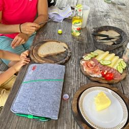 Holztisch mit bayerischer Brotzeit-Platte mit Käse, Schinken, Gemüse, Brot und grauer Reiseunterlagen-Mappe während einer Wanderpause.