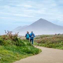 Two hikers in blue jackets walking on coastal path near Moledo with mountain peak in misty background and green vegetation along the trail.