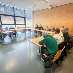 Group of people seated at tables in a modern training room with large windows. An instructor sits at the front conducting a driving safety session.