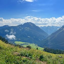 Alpenpanorama von der Jöchelspitze am Lechweg mit grünen Wiesen, Seilbahnseilen und Berggipfeln unter blauem Himmel mit Wolken.
