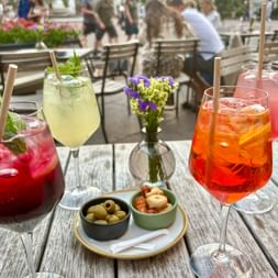 Four colorful cocktails with ice and straws on a wooden table, accompanied by olives and dried tomatoes. A small flower vase decorates the center.