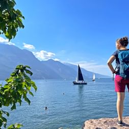 Female hiker with backpack standing on stone shore of Lake Garda, watching sailboats on blue water with mountains in background.