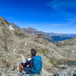 Hiker with blue backpack overlooking Stettiner Hütte on Meraner Höhenweg. Rocky alpine terrain with mountain lake and snow-capped peaks.