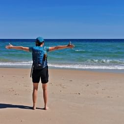 Hiker with backpack and blue cap stands with outstretched arms on sandy beach at Ilha de Tavira, facing turquoise ocean under clear sky.