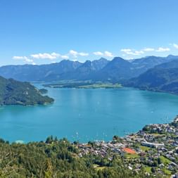Aerial view of turquoise Wolfgangsee surrounded by mountains in Salzkammergut, Austria. A lakeside town and green valleys are visible under blue sky.