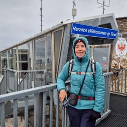 Lächelnde Wanderin in türkisfarbener Jacke an der Zugspitz-Seilbahnstation mit 'Herzlich Willkommen in Tirol'-Schild und Tiroler Wappen.