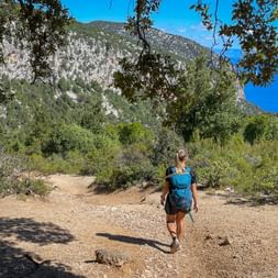 Wanderin mit blauem Rucksack auf felsigem Weg in Norddalmatien, umgeben von grüner Vegetation und felsigen Bergen unter blauem Himmel.