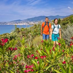 Two hikers with a pink blooming orleander in the foreground and the San Remo flower rivera in the background