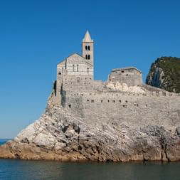 Chiesa di San Pietro in Porto Venere, eine gestreifte Steinkirche mit Glockenturm auf einem Felsvorsprung, umgeben von blauem Meer und grünen Hügeln.