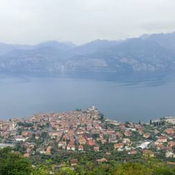 Erhöhter Blick auf Malcesine auf einer Halbinsel im Gardasee, mit rotgedeckten Gebäuden und Kirchturm, umgeben von blauem Wasser und Bergen.