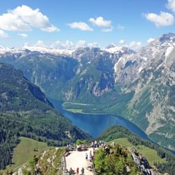 Wanderer auf einem Bergaussichtspunkt mit Blick auf den Königssee, umgeben von schneebedeckten Alpengipfeln und bewaldeten Tälern.