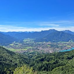 Luftaufnahme des Tegernseer Tals in Bayern mit bewaldeten Hügeln, türkisfarbenem See, grünen Wiesen und Bergketten unter blauem Himmel.