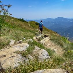 Hiker with blue backpack walking on rocky mountain trail in the Julian Alps, Slovenia, with panoramic view of distant mountain ranges.