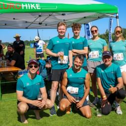 Group of eight runners in turquoise Eurohike shirts posing under green tent at outdoor running event, wearing race numbers on sunny day.