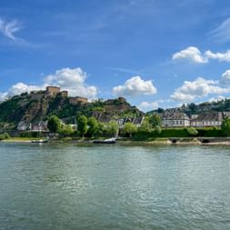 Ehrenbreitstein Fortress on a hilltop overlooking the Rhine River in Koblenz. Historic buildings line the riverbank under a blue sky with white clouds.