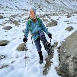 Lächelnde Frau beim Winterwandern mit Wanderstöcken und Rucksack auf verschneitem Berggelände mit großen Felsen in der Landschaft.