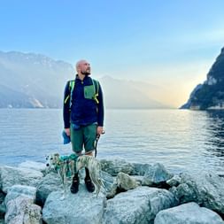 Mann mit Rucksack und kleinem Hund auf felsigem Ufer am Gardasee, mit Bergen und ruhigem Wasser im Hintergrund.