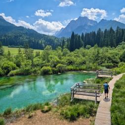 Holzsteg mit Aussichtsplattformen im Naturschutzgebiet Zelenci. Türkises Wasser umgeben von grüner Vegetation, Wald und Bergen unter blauem Himmel.