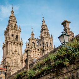 Verzierte barocke Türme der Kathedrale von Santiago de Compostela vor blauem Himmel, mit efeubedeckter Steinmauer und Straßenlaterne im Vordergrund.
