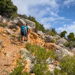 Two hikers with backpacks walking on a rocky mountain trail in Kvarner Bay, Croatia. The path winds through limestone rocks and green vegetation.