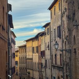 Schmale mittelalterliche Gasse in Volterra mit verwitterten Steingebäuden in Gelb- und Beigetönen, mit Fensterläden und Apothekenschild.