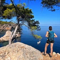 Female hiker with backpack standing on rocky coastal cliff near Deià, viewing deep blue Mediterranean Sea with pine trees and sailboat.