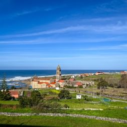 Coastal view of Santa Maria de Oia monastery with bell tower, surrounded by green fields and orange-roofed buildings overlooking the Atlantic Ocean.