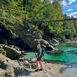 Female hiker with backpack standing on rocky shore of turquoise Soča River in Julian Alps. Suspension bridge spans across river above.