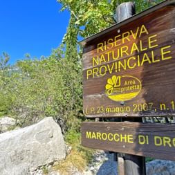 Braunes Holzschild für Riserva Naturale Provinciale Marocche di Dro mit gelbem Text, umgeben von grüner Vegetation und blauem Himmel.