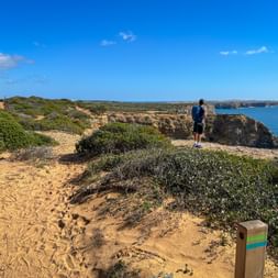 Wanderer mit Rucksack auf sandigem Küstenweg mit Holzwegweiser, Blick auf Klippen und blaues Meer an der Rota Vicentina, Portugal.