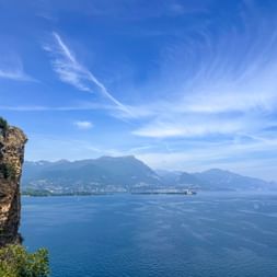 Blick von einer Felsklippe in Manerba del Garda über den blauen Gardasee mit Bergen und Küste in der Ferne unter klarem Himmel.