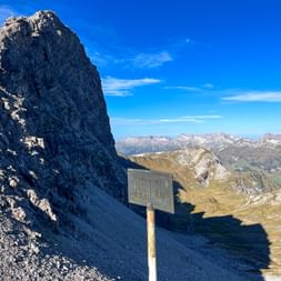 Wooden border marker sign on mountain ridge with dramatic rocky peak and Alpine valley panorama under blue sky in the Allgäu Alps.