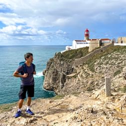 Hiker with backpack on rocky cliff at Cabo São Vicente, Portugal. Red and white lighthouse and fortress visible on cliff top above turquoise ocean.
