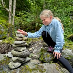 Young girl in light blue hoodie crouching on moss-covered rocks, carefully placing stones on a cairn in a lush green forest near Lake Garda.