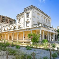 Verlassenes Gebäude im Kolonialstil mit weißen Säulen in Varosha, Zypern. Überwucherte Vegetation umgibt die Struktur, dahinter ein verfallenes Hotel.