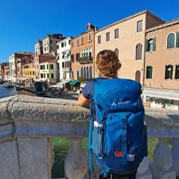 Person mit blauem Eurohike-Rucksack steht auf Steinbrücke in Venedig und betrachtet bunte historische Gebäude entlang des Kanals mit Booten.
