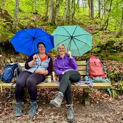 Two female hikers sitting on a wooden bench in a lush green forest, holding blue and turquoise umbrellas, with backpacks beside them.