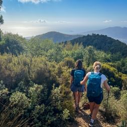 Two hikers with blue backpacks descending a trail on Monte Croce, with forested hills and distant mountains under a blue sky.
