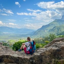 Female hiker with blue backpack sitting on rocky outcrop overlooking green valley in South Tyrol with mountains and blue sky.
