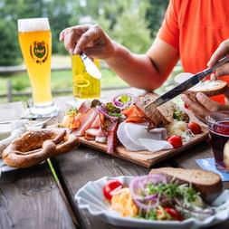 Person im orangenen Shirt genießt traditionelle bayerische Brotzeit im Freien mit Bierglas, Holzbrett mit Aufschnitt, Käse und Brot.