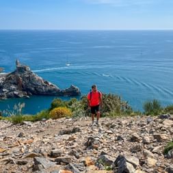 Hiker in red jacket on rocky trail overlooking Porto Venere bay with church on promontory, blue Mediterranean Sea, and sailboats.