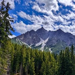 Felsiger Serles-Gipfel in den Stubaier Alpen mit Schneeresten, umgeben von dichtem grünen Nadelwald unter blauem Himmel mit weißen Wolken.