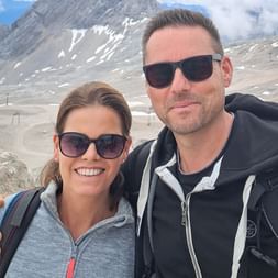 Smiling couple with sunglasses and backpacks taking a selfie in the Zugspitze mountain region with rocky peaks and snow patches visible.