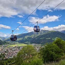Zwei blaue Gondeln über Sterzing mit Bergpanorama. Die Stadt liegt in einem Tal, umgeben von grünen Alpengipfeln unter bewölktem Himmel.