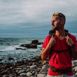 Mann im roten Shirt mit Rucksack und Sonnenbrille am Kieselstrand von Hovs Hallar, Schweden, mit felsiger Küste und Meer im Hintergrund.