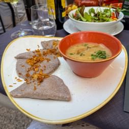 Two buckwheat dumplings with crispy onions and creamy soup in orange bowl. Fresh salad and wine glasses visible in background.